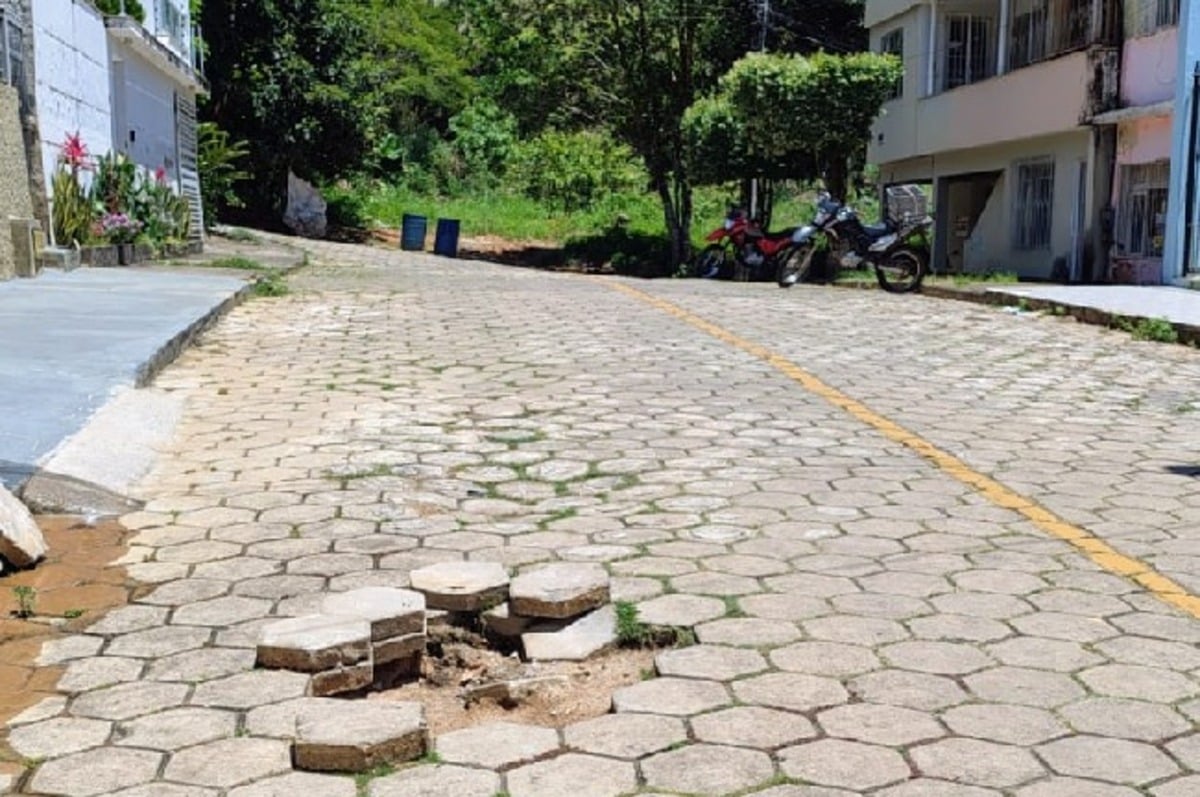 Buraco aberto em uma rua pavimentada com blocos de concreto, com várias peças soltas e empilhadas ao redor da abertura. A via está em área residencial, com casas à direita e vegetação ao fundo. Algumas motocicletas estão estacionadas à sombra de árvores mais à frente.