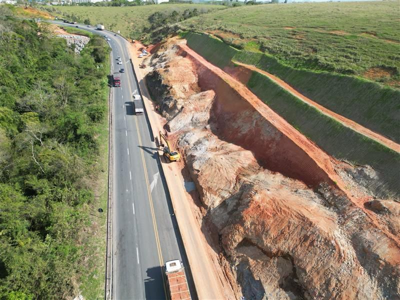 Vista aérea de um trecho da BR-101 onde máquinas escavadeiras trabalham no desmonte de rochas às margens da rodovia. À esquerda, veículos circulam normalmente pela pista, enquanto à direita é possível ver a encosta terraplanada e grandes cortes no solo como parte das obras de duplicação.