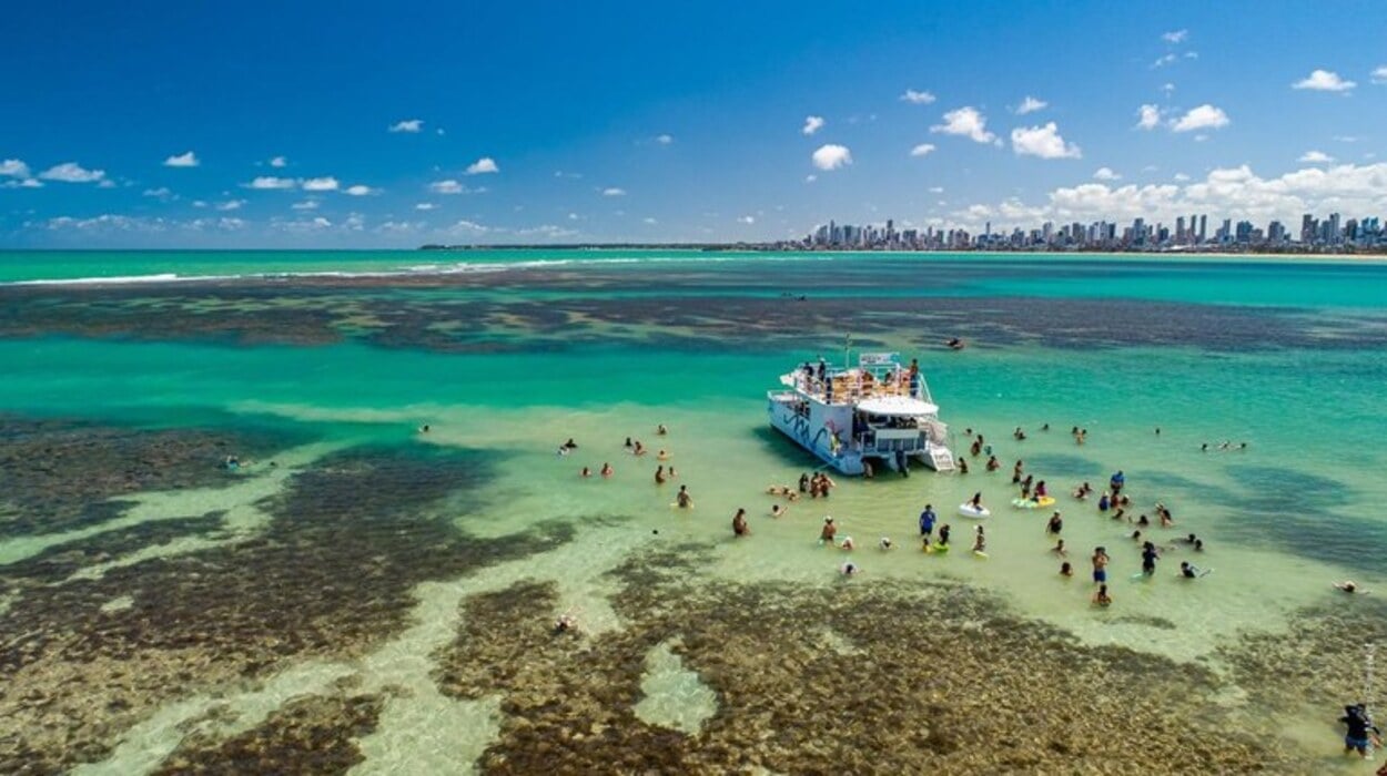 Turistas nadam em águas rasas e cristalinas próximas a um catamarã ancorado em uma área de recifes, com o mar em tons de verde e azul e a skyline de João Pessoa ao fundo sob céu ensolarado.