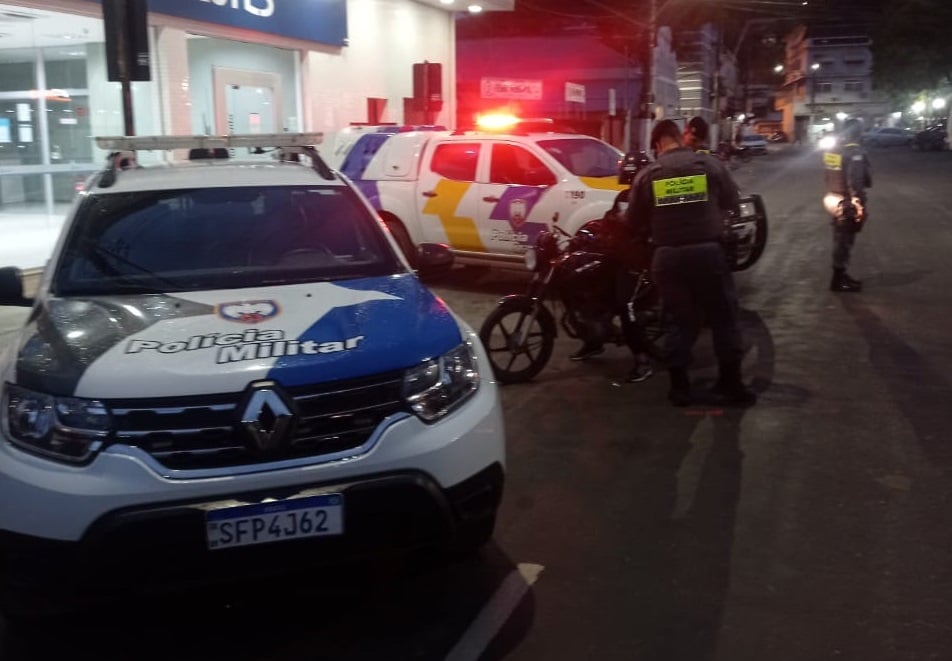 Viaturas da Polícia Militar estão paradas em frente a uma agência bancária durante operação noturna. Policiais abordam um motociclista enquanto outro agente realiza fiscalização próxima, com giroflex ligado ao fundo.