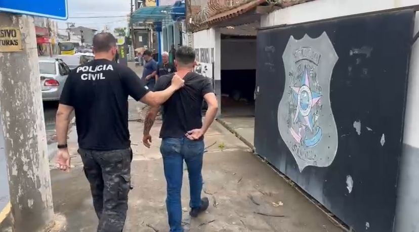 Imagem mostra um policial civil conduzindo um homem algemado pelas costas até a entrada de uma delegacia. O agente segura o suspeito pelo ombro enquanto ambos caminham por uma calçada. Ao fundo, é possível ver o portão da unidade policial com o brasão da instituição e alguns veículos estacionados na rua.