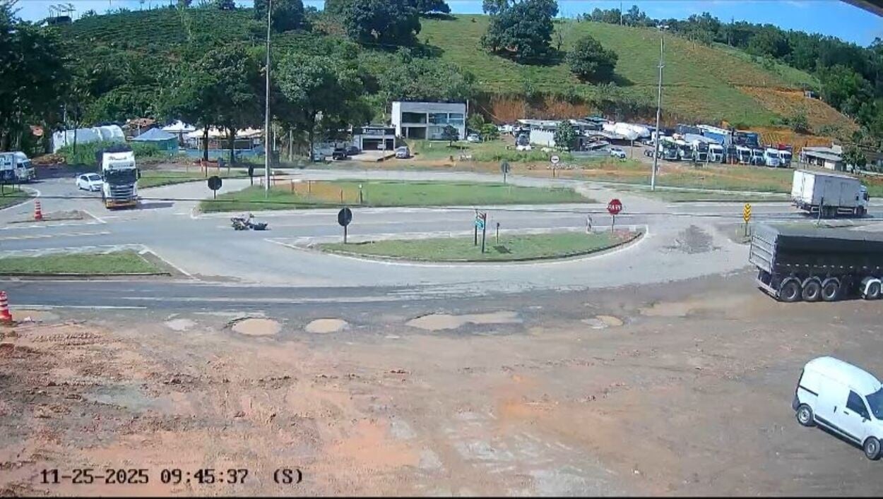 Imagem de câmera de segurança mostra o trevo de Piúma, no km 370 Sul da BR-101. No registro, uma motocicleta aparece caída no asfalto após um acidente, enquanto um caminhão segue pela via. A cena revela o cruzamento amplo, com veículos passando ao redor e área de estacionamento de caminhões ao fundo.
