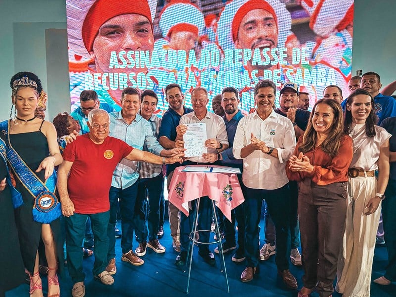 Grupo de autoridades e representantes das escolas de samba posa sorrindo após a assinatura do repasse de recursos para o carnaval 2026; governador Renato Casagrande exibe o documento no centro da foto, cercado por apoiadores no palco decorado com imagens festivas.