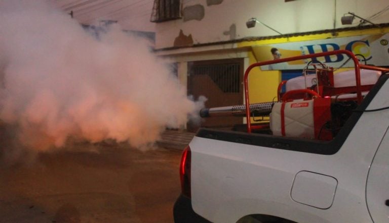 a imagem mostra um carro espalhando fumaça contra os mosquitos próximos de casas
