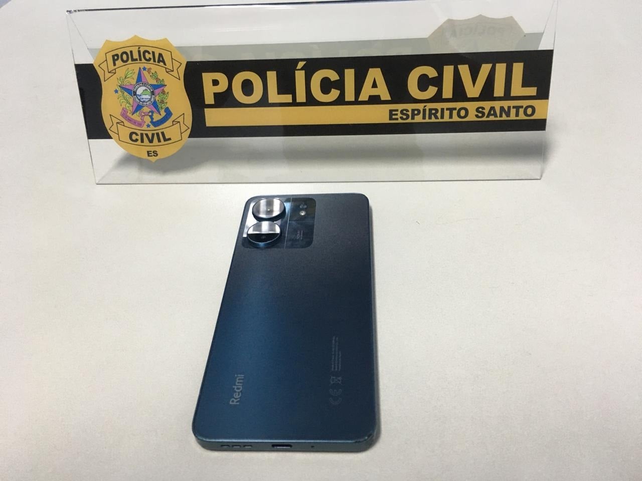 Celular preto sobre uma mesa branca, com uma placa da Polícia Civil.