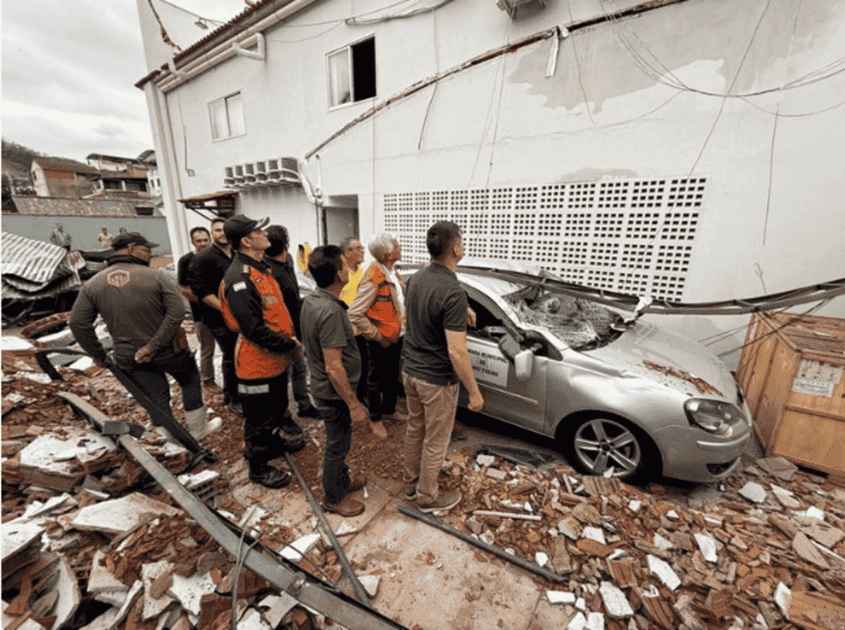 Um grupo de agentes da Defesa Civil e outras pessoas observa os estragos causados pelo desabamento de parte de uma estrutura sobre um carro prata. O veículo está gravemente danificado, coberto por destroços de tijolos e telhas. A cena ocorre ao lado de um prédio, cuja parede também apresenta danos e fiações caídas. O chão está repleto de entulhos.