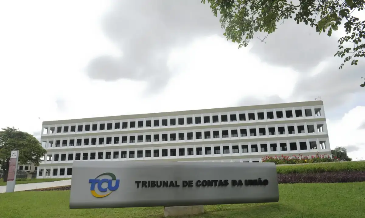 Fachada do prédio do Tribunal de Contas da União, em Brasília, vista de frente, com estrutura branca e várias janelas alinhadas; em primeiro plano há uma placa cinza metálica com o logotipo do TCU sobre um gramado verde, sob céu nublado.