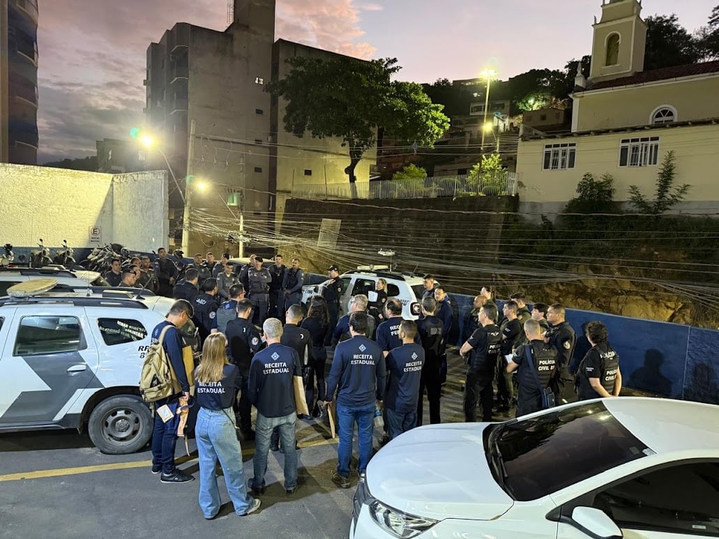 Grupo numeroso de agentes da Receita Estadual, Polícia Civil e Polícia Militar se reúne em um pátio ao amanhecer. Eles usam uniformes identificados e estão ao lado de viaturas, enquanto recebem orientações para uma operação conjunta. Ao fundo, prédios e uma igreja compõem o cenário urbano.