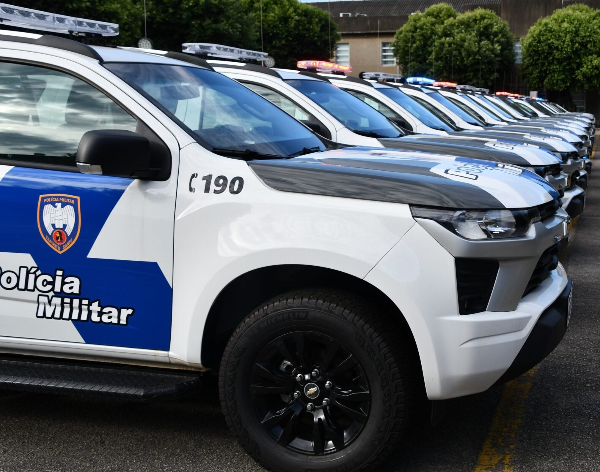 Imagem consta a lateral de uma viatura da Polícia Militar, branca e azul.
