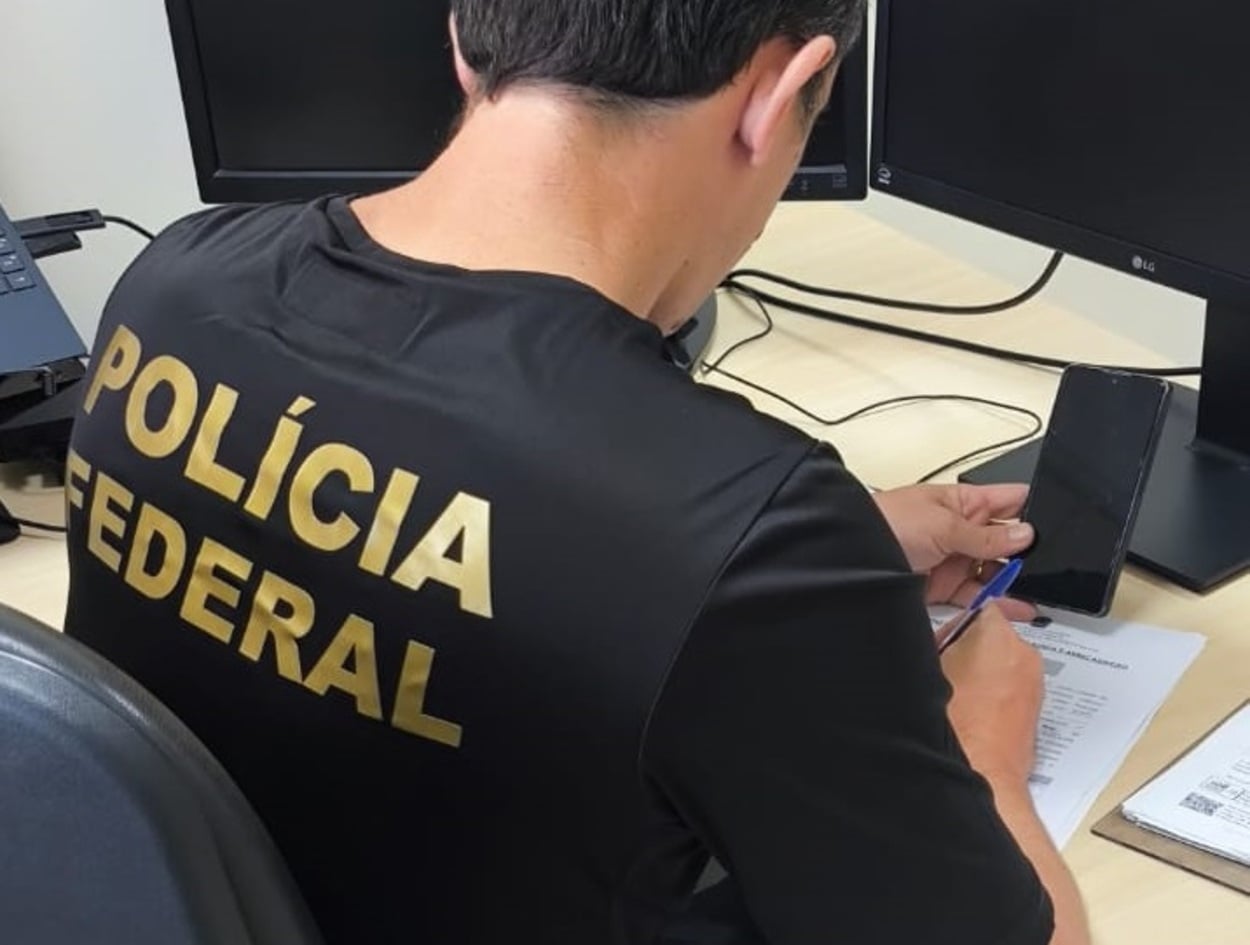 Operação da PF - Polícia federal sentando em uma mesa analisando documentos, com um celular na mão. - Operação da PF