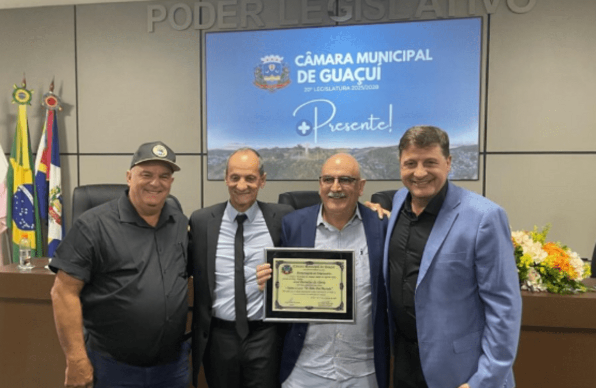 Quatro homens posam sorrindo em uma cerimônia na Câmara Municipal de Guaçuí. Um deles, ao centro, segura uma placa de homenagem s funerária Em Vida. Ao fundo, há um telão com o brasão do município e os dizeres 'Câmara Municipal de Guaçuí – Presente!'. Bandeiras oficiais e arranjos de flores também compõem o ambiente formal.