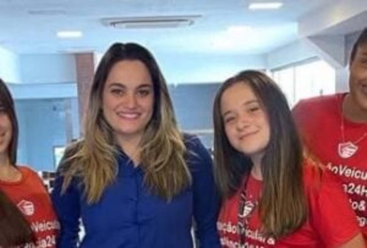 Foto mostra duas jovens, em pé, com uniformes de trabalho azul e vermelho.