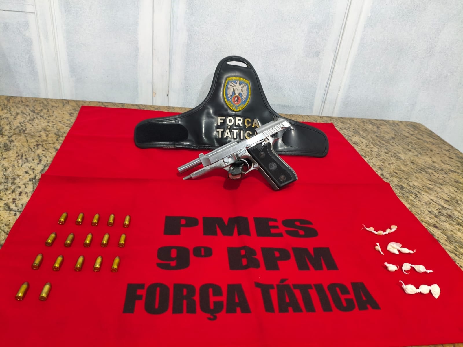 Mesa de granito exibe uma pistola prateada, um carregador e várias munições organizadas ao lado de pequenos papelotes de drogas. Os itens estão sobre um pano vermelho com a inscrição “PMES 9º BPM Força Tática”, acompanhado de um suporte preto com o brasão da Polícia Militar.