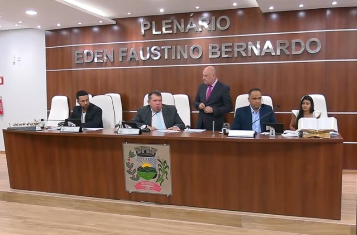 Plenário Eden Faustino Bernardo durante sessão oficial. Cinco pessoas estão sentadas atrás da mesa diretora em cadeiras brancas. No centro, o presidente da sessão fala ao microfone. À esquerda e à direita, outros membros acompanham a reunião com documentos à mesa. Uma mulher está à ponta direita com um livro aberto à sua frente. O brasão do município está fixado na parte frontal da mesa.