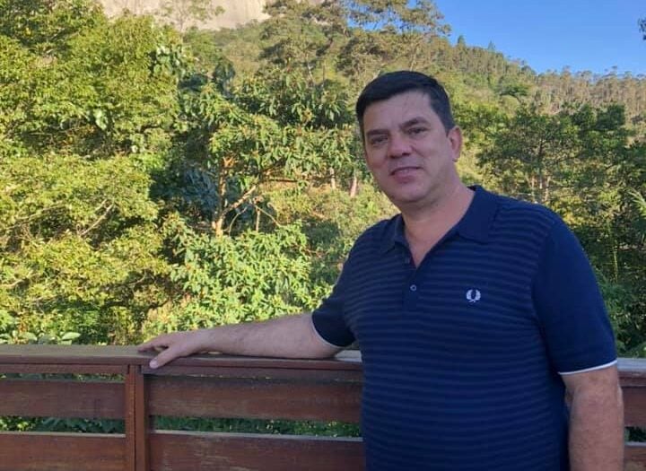 Homem posa para foto encostado em uma cerca de madeira, com vegetação densa ao fundo. Ao fundo, aparece uma grande formação rochosa iluminada pelo sol, em um dia de céu azul.