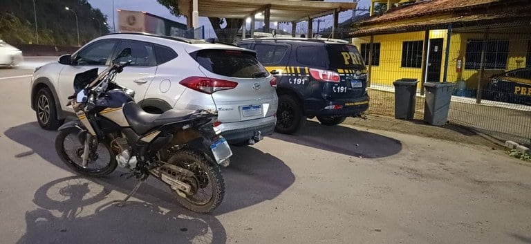 Imagem mostra uma motocicleta e um Hyundai Tucson apreendidos pela PRF estacionados ao lado de uma viatura da Polícia Rodoviária Federal em frente ao posto policial em Viana, durante a noite.
