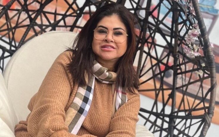 Foto retrata mulher, sentada em uma poltrona, com casado marrom e chale branco.