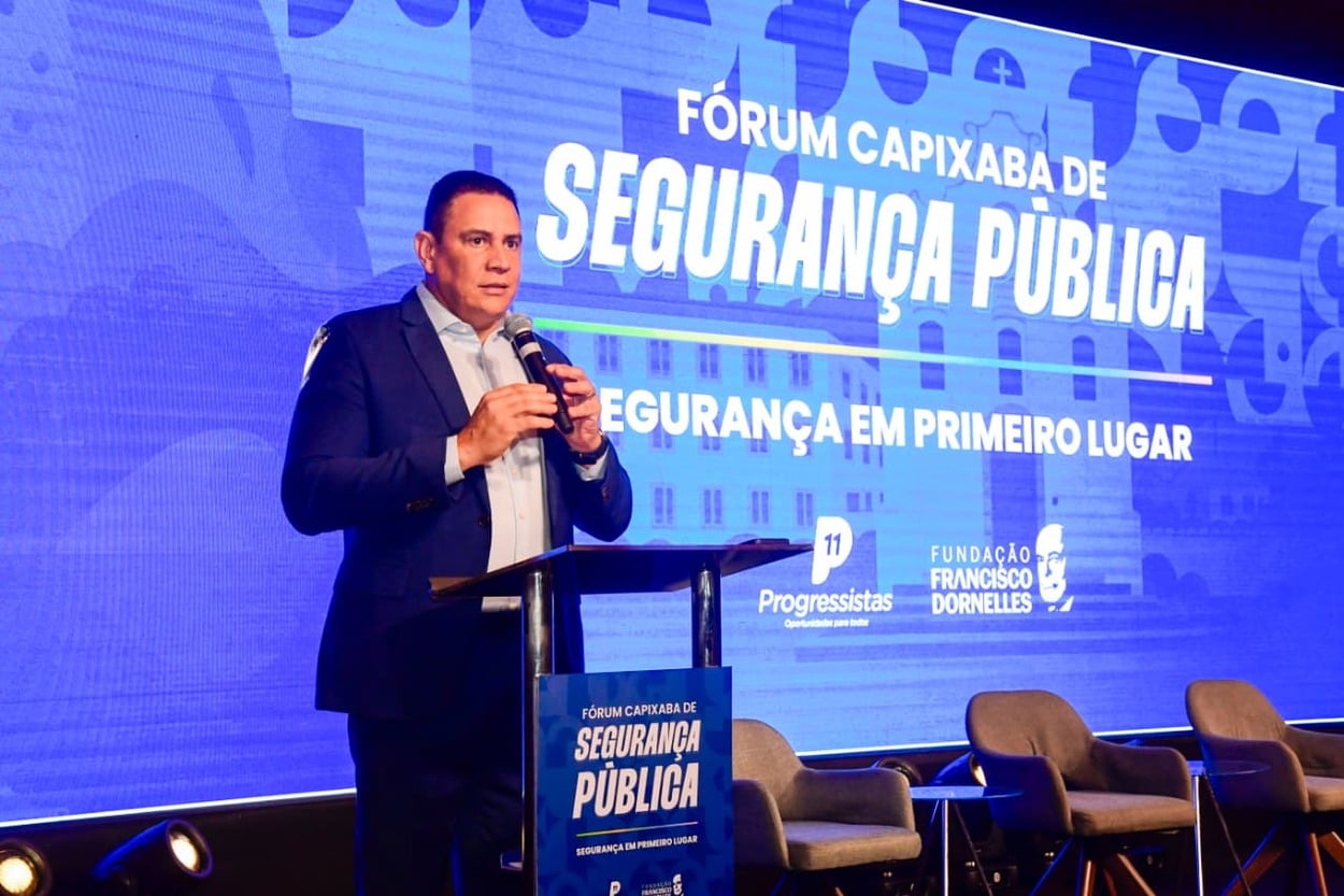 Um homem vestido com terno azul e camisa clara fala ao microfone em um púlpito durante um evento. Atrás dele, um grande painel azul exibe os dizeres “Fórum Capixaba de Segurança Pública” e “Segurança em primeiro lugar”, além de logotipos do partido Progressistas e da Fundação Francisco Dornelles. No palco, ao lado do orador, há cadeiras vazias arrumadas para o evento. A iluminação destaca o palestrante e o fundo.