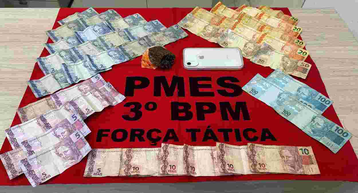 Cédulas de dinheiro, um celular e uma porção de droga apreendidos pela Força Tática do 3º BPM em Jerônimo Monteiro, expostos sobre um pano vermelho da PMES.