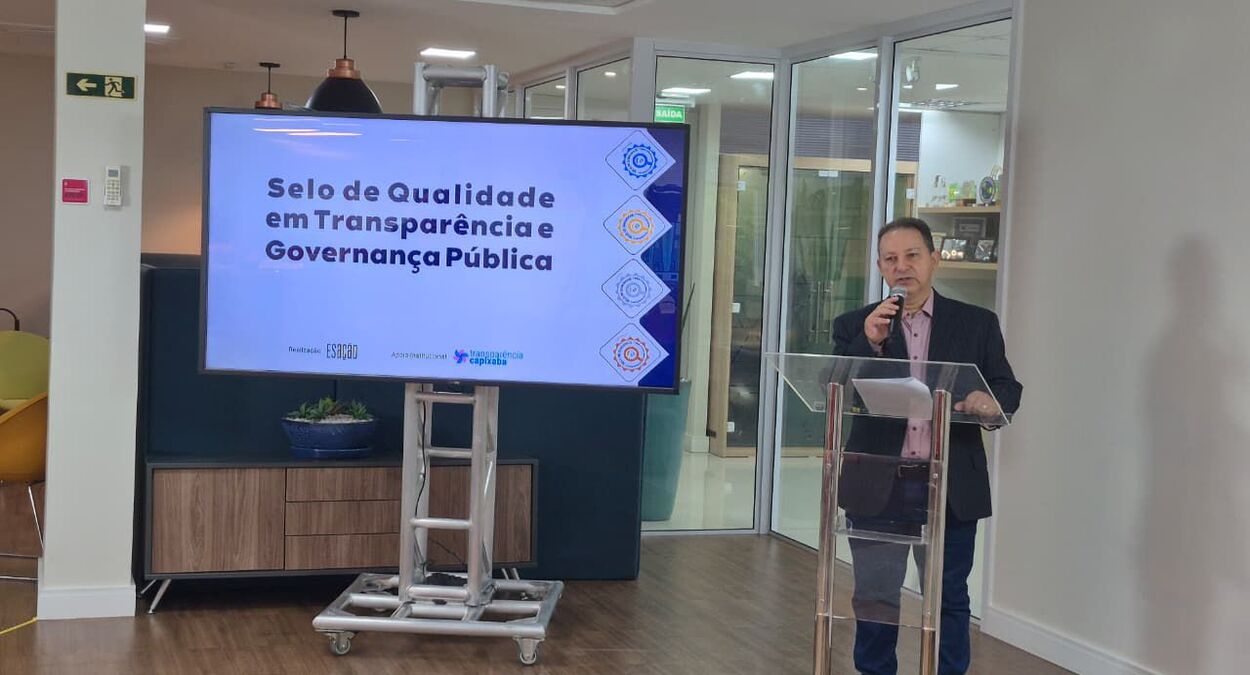 Homem apresenta o Selo de Qualidade em Transparência 2025 durante cerimônia, ao lado de tela que exibe o nome do programa em ambiente institucional.