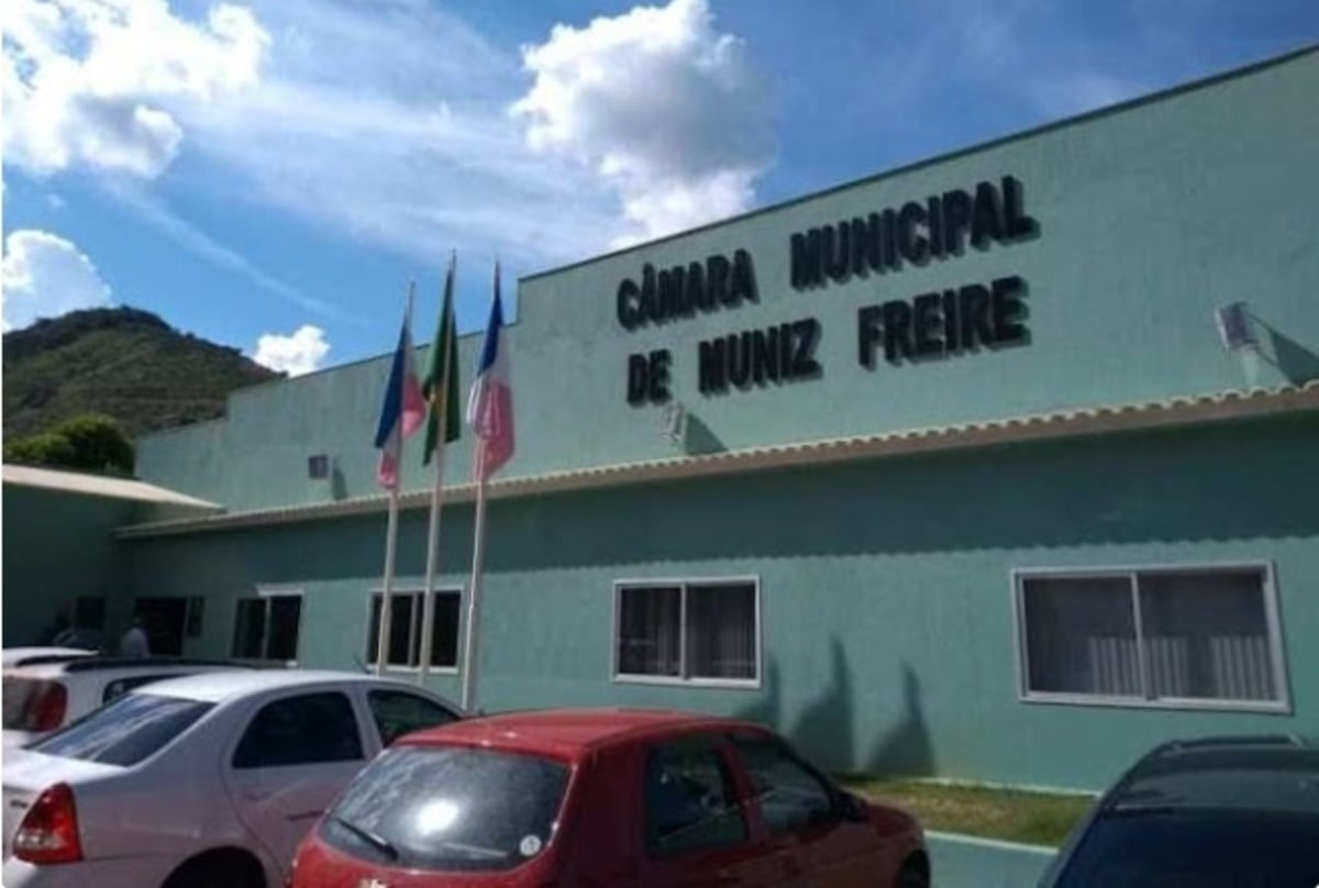 Fachada da Câmara Municipal de Muniz Freire em dia ensolarado, com prédio verde-claro, letreiro em destaque na parte superior, quatro mastros com bandeiras hasteadas e carros estacionados em frente ao edifício.