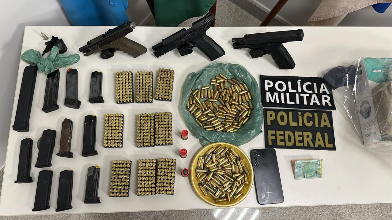 Mesa exibe materiais apreendidos na Operação Derivada: três pistolas, diversos carregadores, centenas de munições de vários calibres distribuídas em bandejas e suportes, cartuchos avulsos, celular, dinheiro e identificações da Polícia Militar e da Polícia Federal.