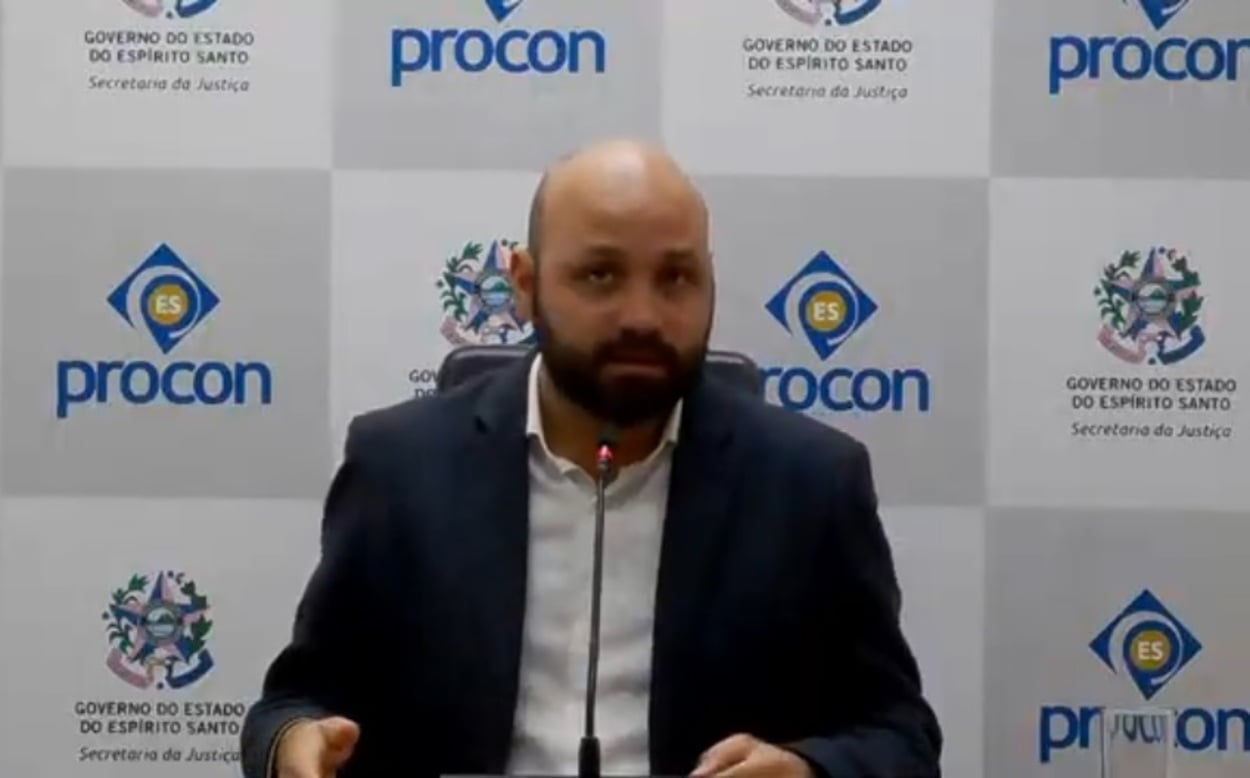 Homem sentado em uma mesa, usando terno escuro e camisa branca, falando diante de um microfone. Ao fundo, há um painel institucional com logotipos do Procon-ES e do Governo do Estado do Espírito Santo.