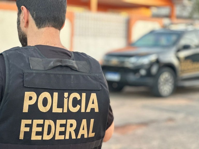 Imagem retrata policial federal com colete ao lado de viatura.