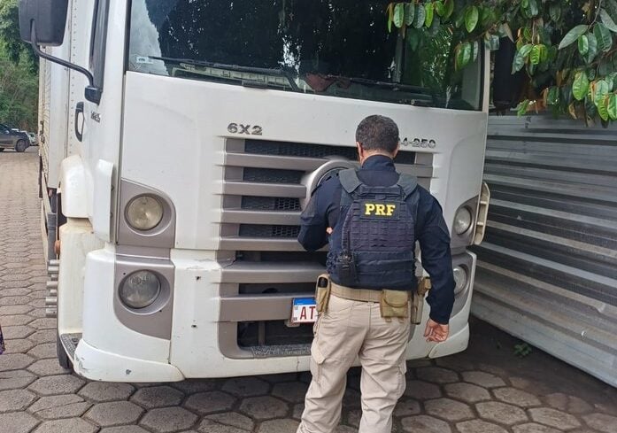 Imagem mostra caminhão roubado recuperado pela PRF e um policial em pé na frente do veículo.