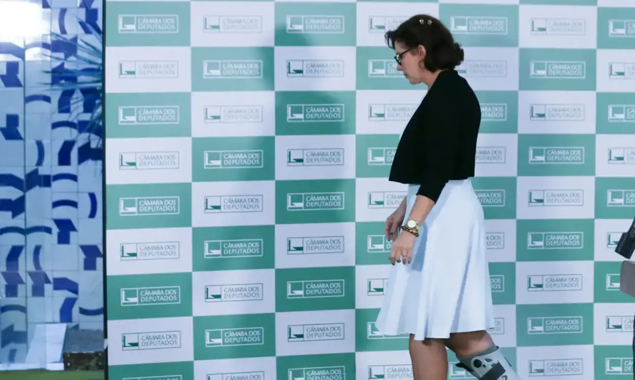 Uma mulher caminha em frente a um painel institucional da Câmara dos Deputados, composto por quadros verdes e brancos com o logotipo da Casa. Ela veste saia branca, blusa escura e usa uma bota ortopédica na perna direita. A cena parece ocorrer em um ambiente interno iluminado.