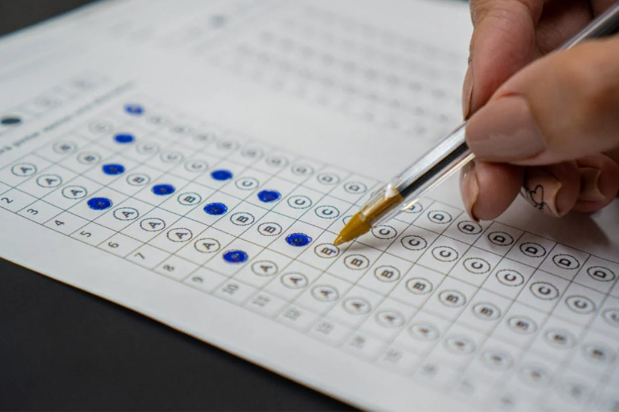 Uma mão segura uma caneta esferográfica enquanto marca respostas em um cartão de prova de concurso. O cartão contém linhas de alternativas com letras de A a E, e vários círculos já estão preenchidos com tinta azul. O foco está na caneta tocando o papel, sugerindo que a pessoa está no processo de responder às questões.