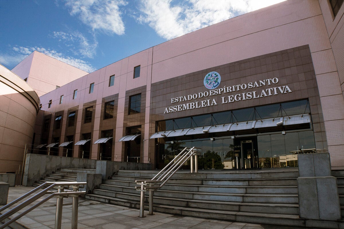 Fachada da Assembleia Legislativa do Estado do Espírito Santo, com escadaria ampla à frente, corrimãos metálicos e a entrada principal com portas de vidro sob marquise. O prédio possui revestimento em tons de rosa e bege, com céu azul ao fundo.