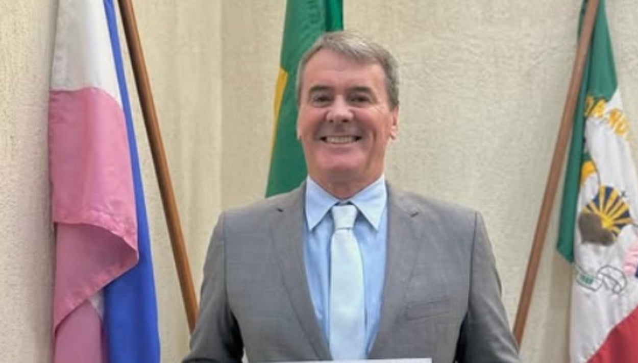 Homem de terno cinza e gravata clara sorri enquanto posa segurando um documento. Ele está em ambiente institucional, com bandeiras ao fundo, incluindo as cores verde e amarela.