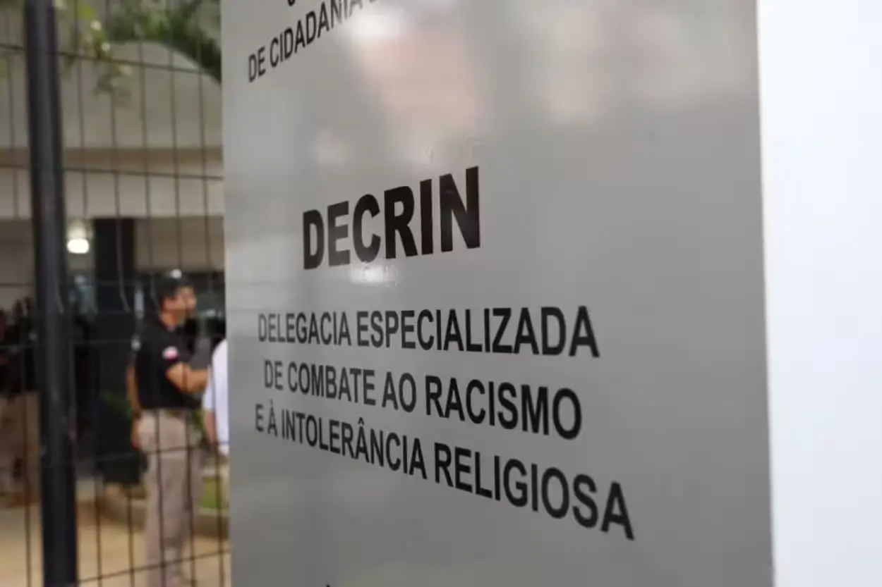 Placa metálica da Decrin, Delegacia Especializada de Combate ao Racismo e à Intolerância Religiosa, com o nome da unidade em destaque. Ao fundo, desfocado, aparecem agentes uniformizados e parte da área externa do prédio, vista através de uma grade.