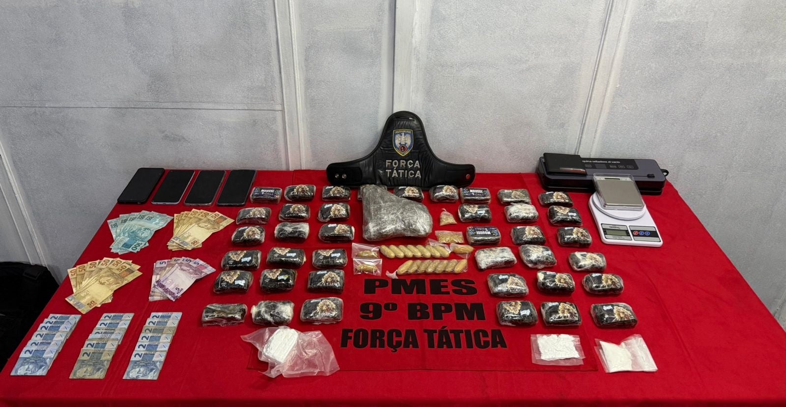 Mesa da Força Tática exibindo grande apreensão de drogas em Cachoeiro, com porções de entorpecentes, dinheiro, celulares, balanças de precisão e material de embalagem organizados sobre um pano vermelho.