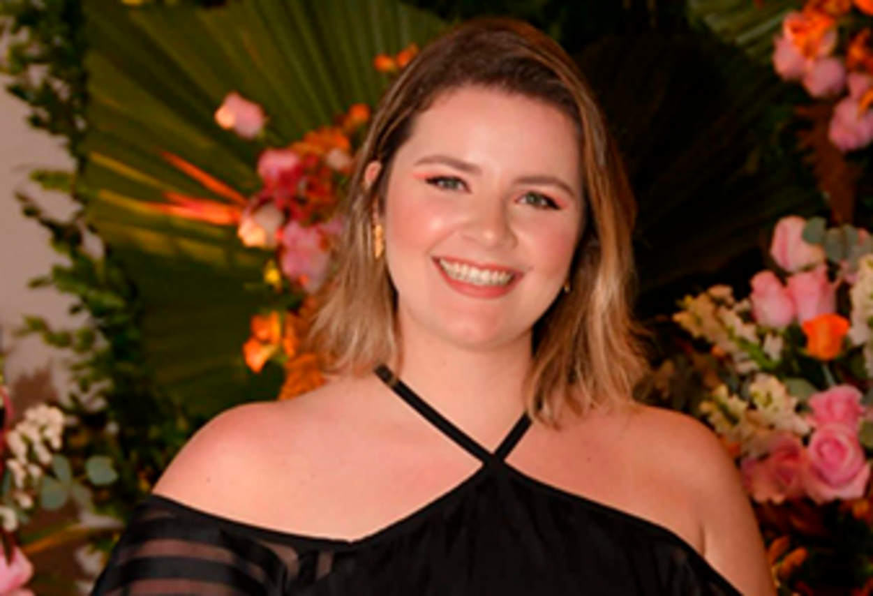 Imagem retrata jovem loira, usando vestido presto, sorrindo, próximo de flores.