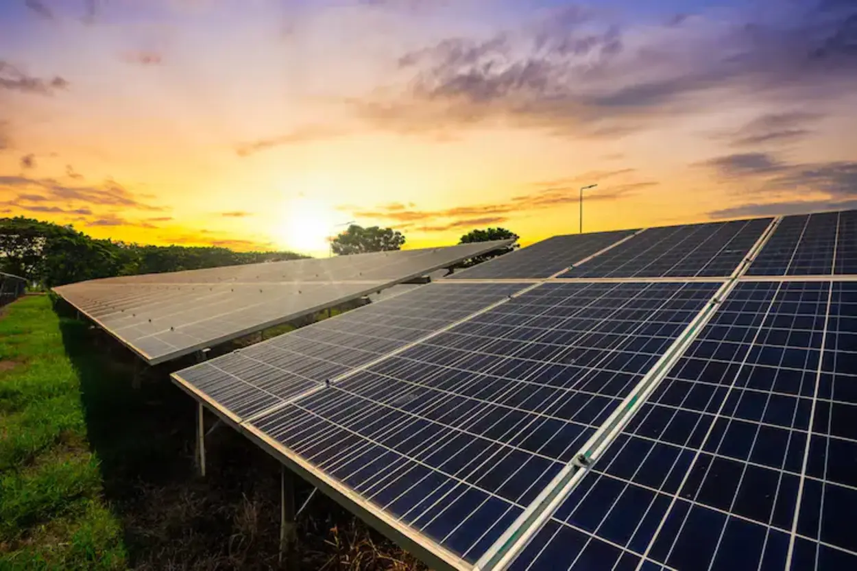 Painéis solares instalados em uma área verde, alinhados em fileiras, recebendo a luz do sol durante o pôr do sol, com céu amarelado e árvores ao fundo.