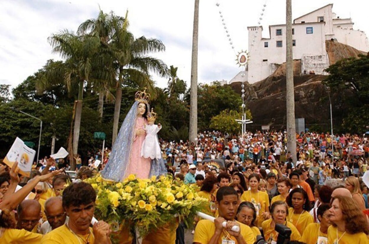 Procissão da Festa da Penha em Vila Velha, com multidão acompanhando a imagem de Nossa Senhora da Penha carregada por fiéis diante do Convento da Penha ao fundo.