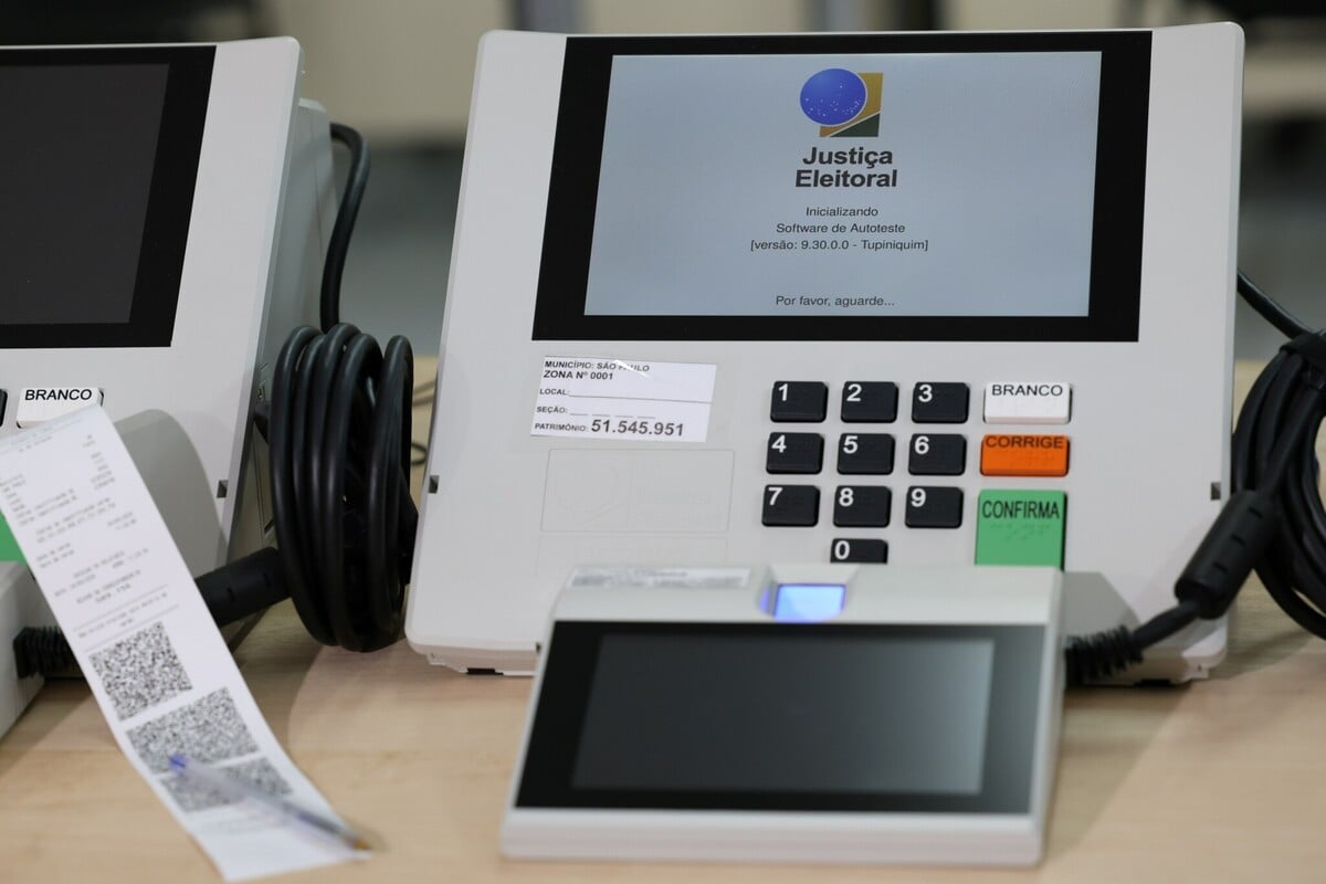 Máquina de votação eletrônica do Tribunal Superior Eleitoral (TSE) sobre uma mesa, exibindo na tela a mensagem de inicialização do sistema. Ao lado do teclado numérico, com botões “Branco”, “Corrige” e “Confirma”, há cabos conectados e um terminal auxiliar com tela sensível ao toque. Em primeiro plano, um comprovante impresso com QR code aparece parcialmente enrolado.