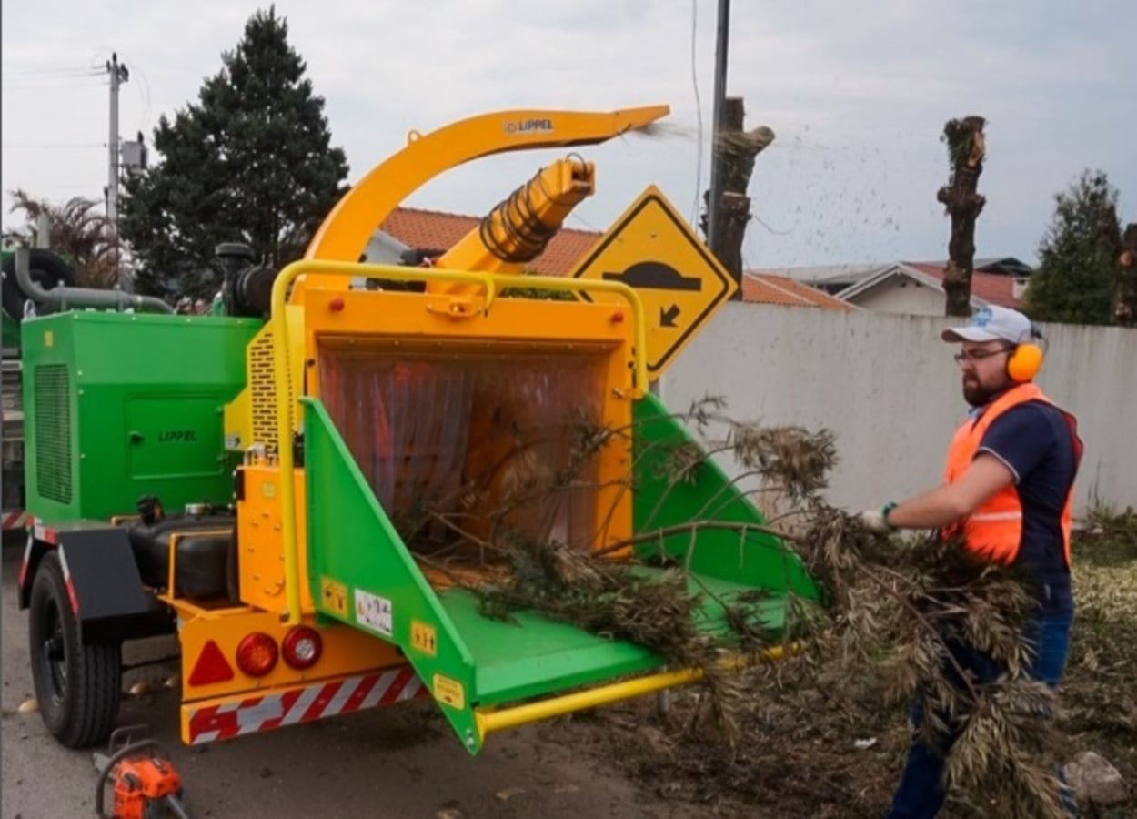 Servidor municipal utiliza triturador de galhos para processar resíduos de poda urbana, com equipamento acoplado a veículo, em via pública residencial.