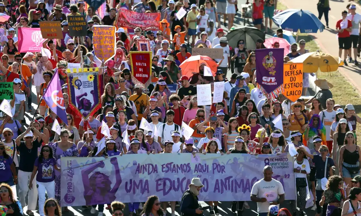 Multidão de mulheres marcha em protesto durante o dia, carregando faixas, cartazes e bandeiras com mensagens em defesa dos direitos das mulheres, educação antimachista e estado laico; grupo avança por avenida sob sol forte, algumas participantes usam bonés, lenços e sombrinhas.