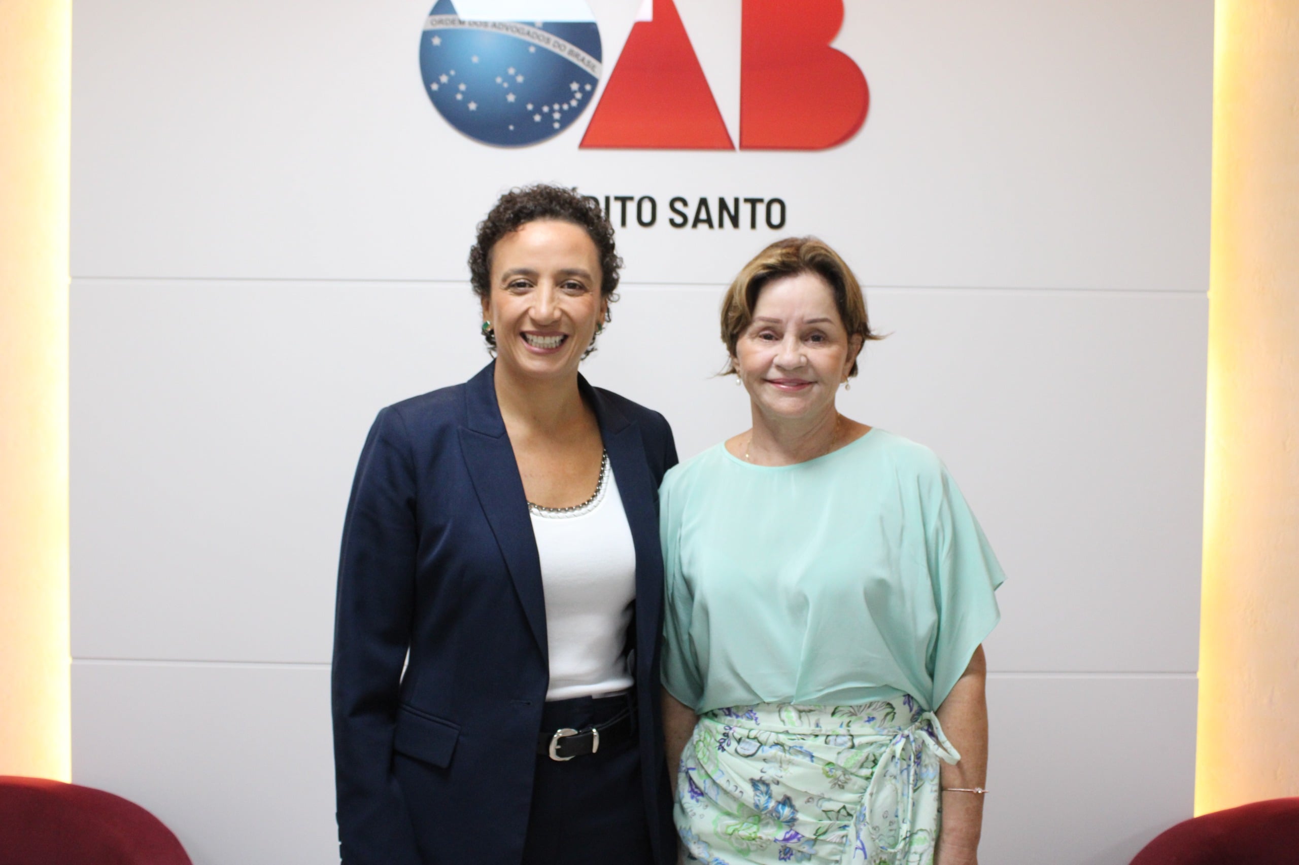 Imagem retrata duas mulheres, atrás, um backdrop com símbolo da OAB