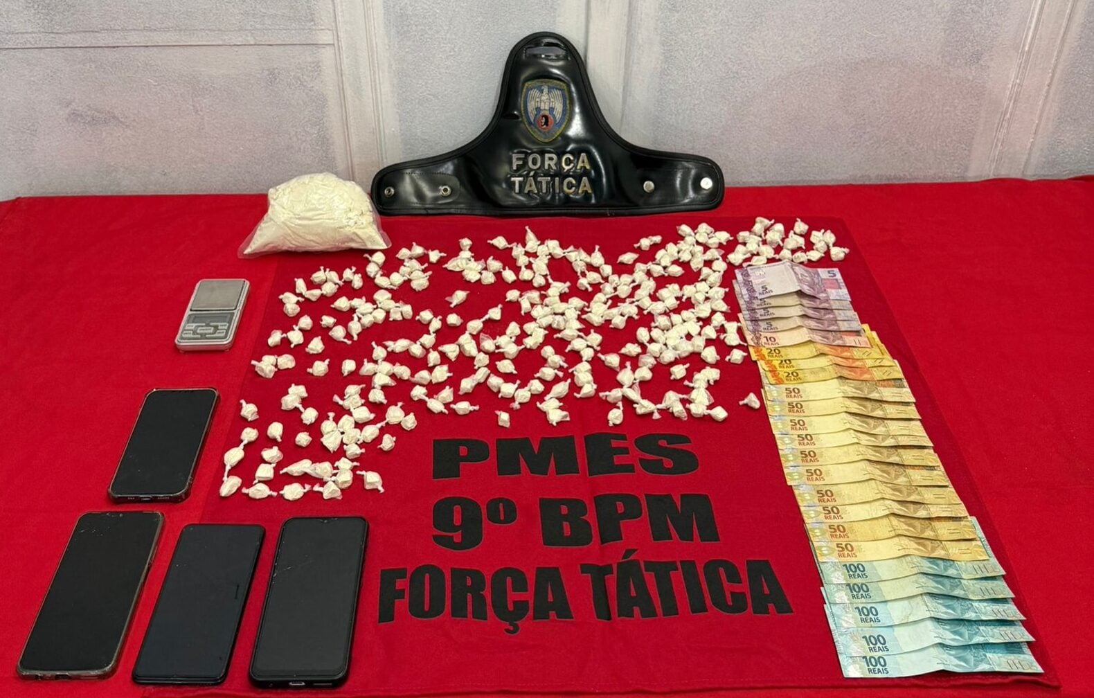Mesa coberta com um pano vermelho da Força Tática do 9º BPM exibindo materiais apreendidos: dezenas de papelotes de cocaína, uma porção maior da droga embalada, uma balança de precisão, quatro celulares e diversas cédulas de dinheiro organizadas em fileiras. Acima dos itens, há um distintivo rígido da Força Tática apoiado na parede.