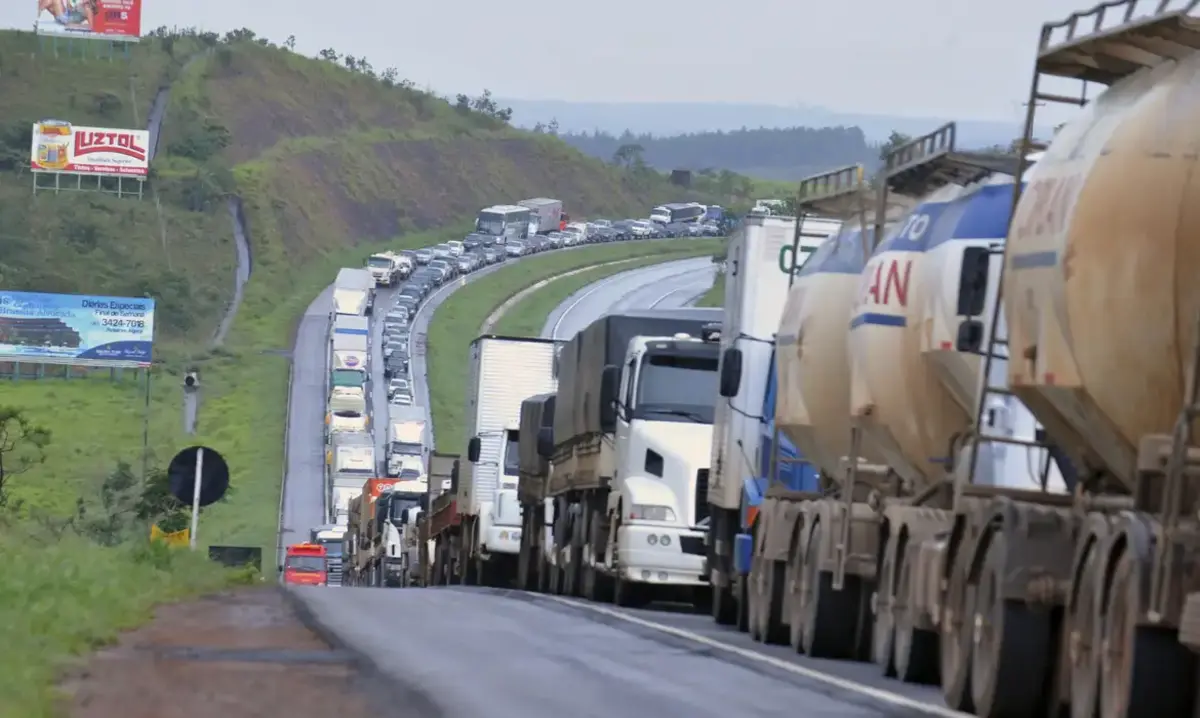 Uma longa fila de caminhões e carros ocupa ambos os sentidos de uma rodovia em região montanhosa. Os veículos estão praticamente parados, formando um congestionamento que se estende até onde a estrada curva e desaparece ao fundo. À direita, vários caminhões-tanque dominam o primeiro plano; à esquerda, outdoors aparecem sobre a encosta coberta de vegetação. O céu está nublado e o asfalto parece úmido, sugerindo chuva recente.