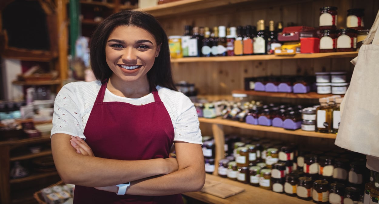 Empreendedora sorrindo em sua loja com prateleiras de produtos ao fundo, representando participantes da Feira do Parceria Legal em Guaçuí.