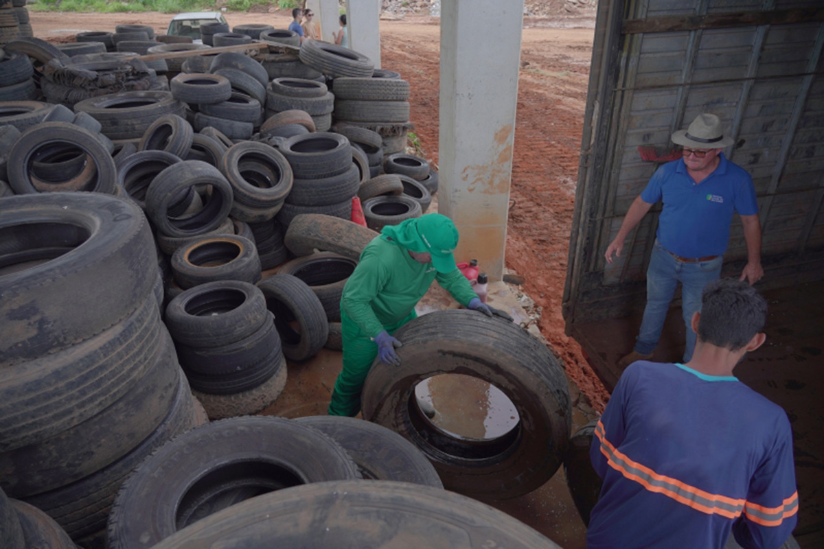 A imagem mostra um grande galpão com dezenas de pneus velhos empilhados no chão. No centro, um trabalhador usando uniforme verde, luvas e boné pega um pneu grande que está cheio de água acumulada. À direita, dois homens observam a operação; um deles usa chapéu e camisa azul, e o outro veste camisa azul marinho com faixas reflexivas. O ambiente ao fundo é de terra vermelha, indicando área externa ou de descarte.