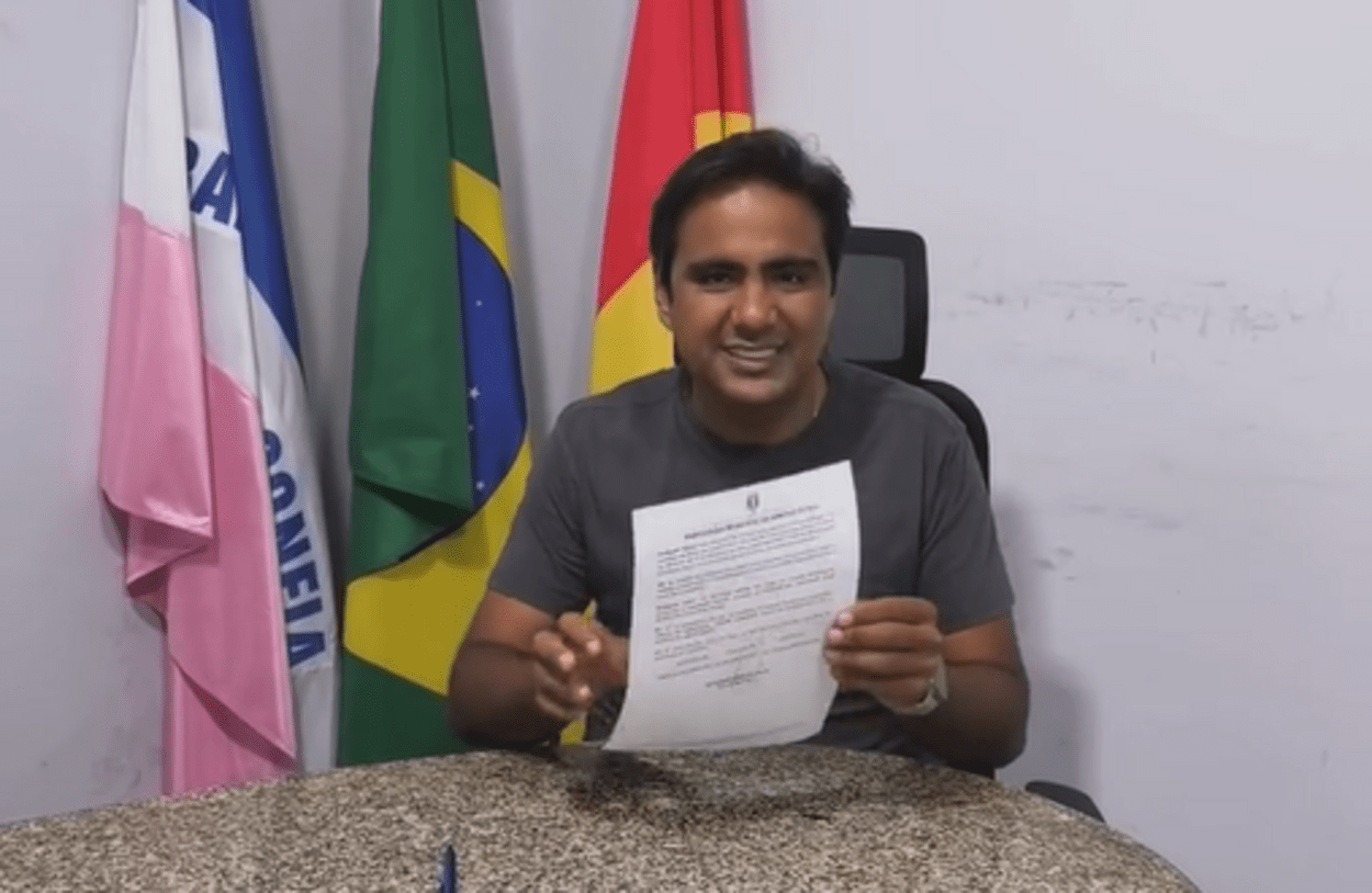 Homem sentado atrás de uma mesa, em ambiente institucional, segurando um documento impresso com as duas mãos e sorrindo para a câmera. Ao fundo, aparecem bandeiras posicionadas em suportes, incluindo a do Brasil e outras bandeiras oficiais. Sobre a mesa há uma caneta, e o cenário sugere um gabinete ou sala administrativa.