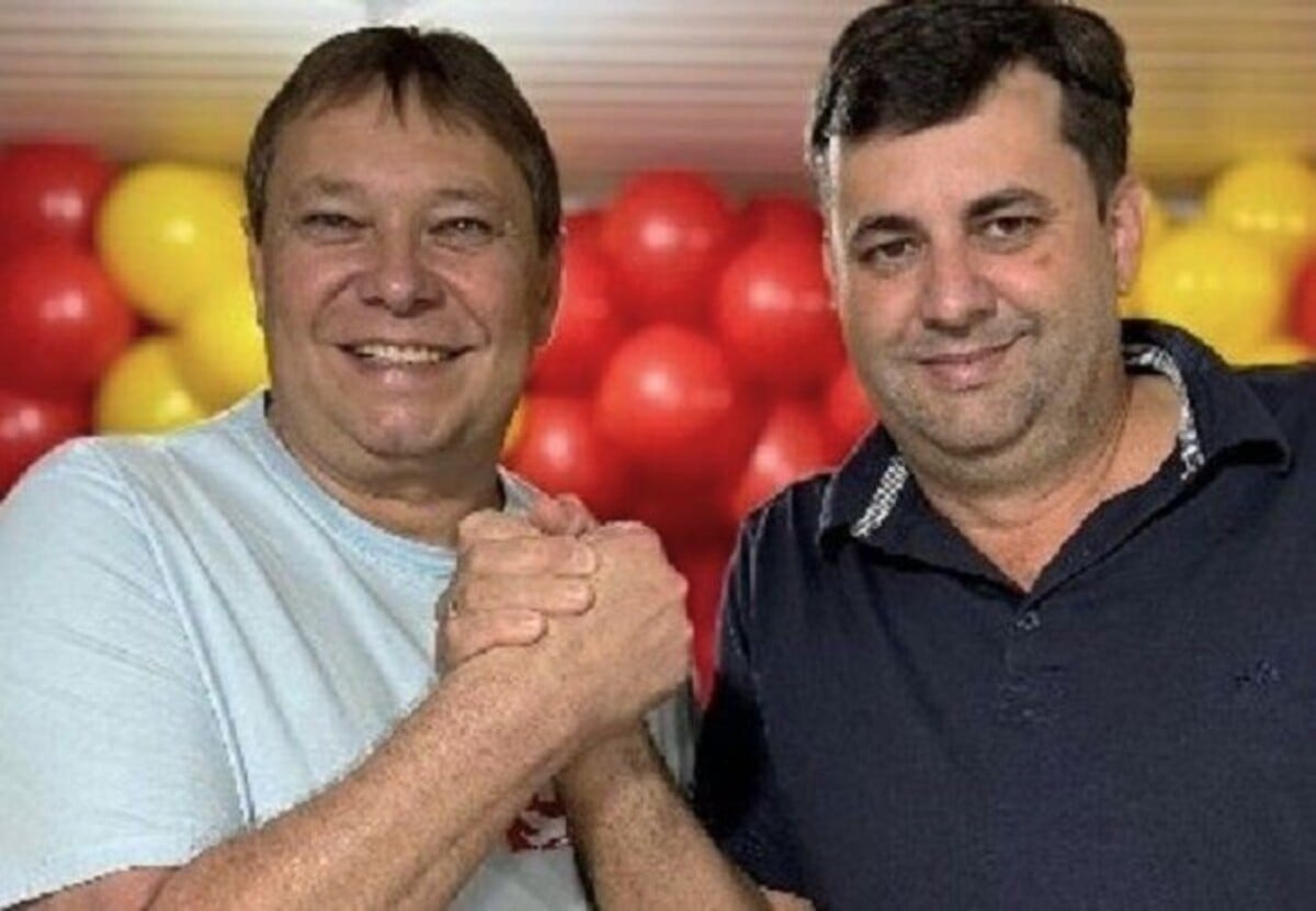 Dois homens posam sorrindo para a foto, apertando as mãos em gesto de cumprimento. Ao fundo, há uma decoração com balões vermelhos e amarelos.