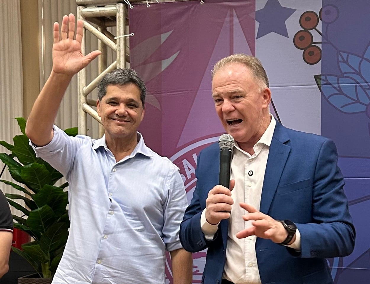 Dois homens em um evento institucional: à esquerda, um homem de camisa clara sorri e acena com a mão levantada; à direita, outro homem de blazer azul segura um microfone e fala ao público, com painel gráfico ao fundo e estrutura metálica no palco.