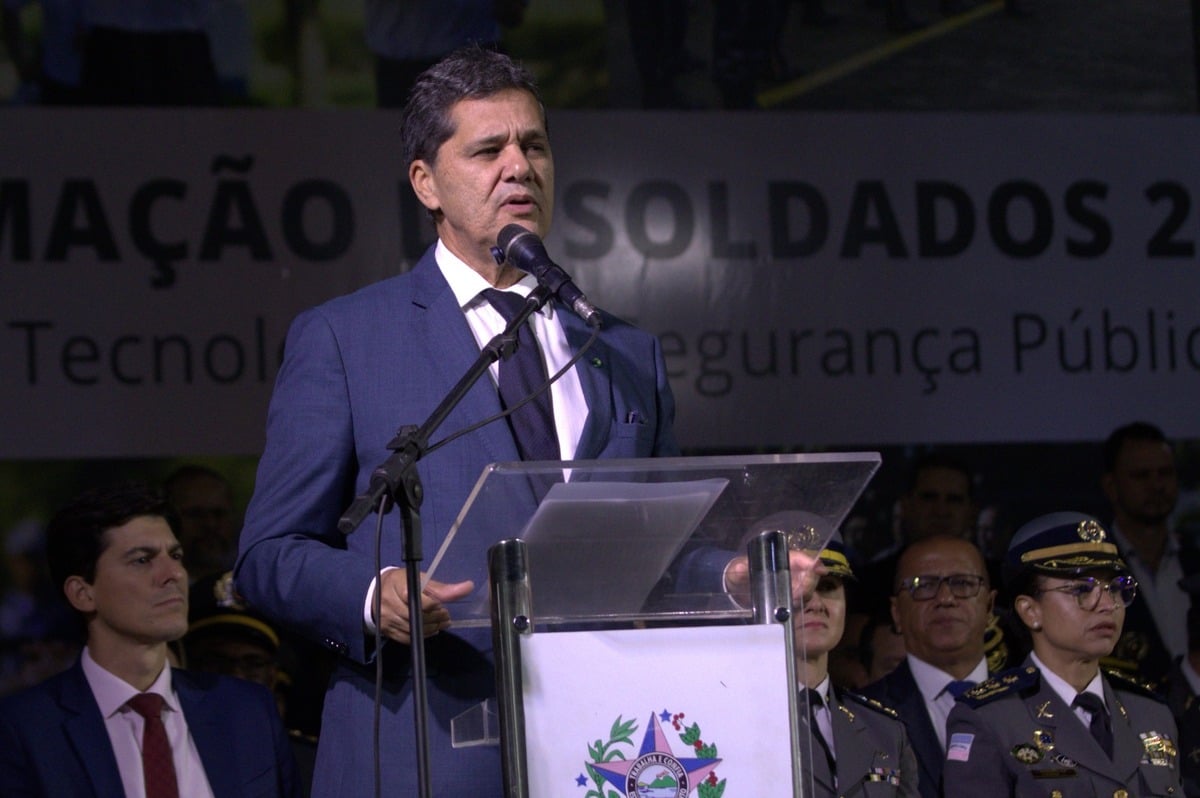 Homem de terno azul fala ao microfone em um púlpito transparente durante cerimônia oficial. Atrás dele, autoridades militares fardadas e outras pessoas acompanham o evento. Um banner ao fundo exibe palavras relacionadas à formação de soldados e à segurança pública.